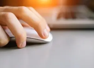 Close-up of a persons hand using a white computer mouse on a gray surface. The background shows part of a keyboard with a warm light glow. The focus is on the fingers and the mouse.