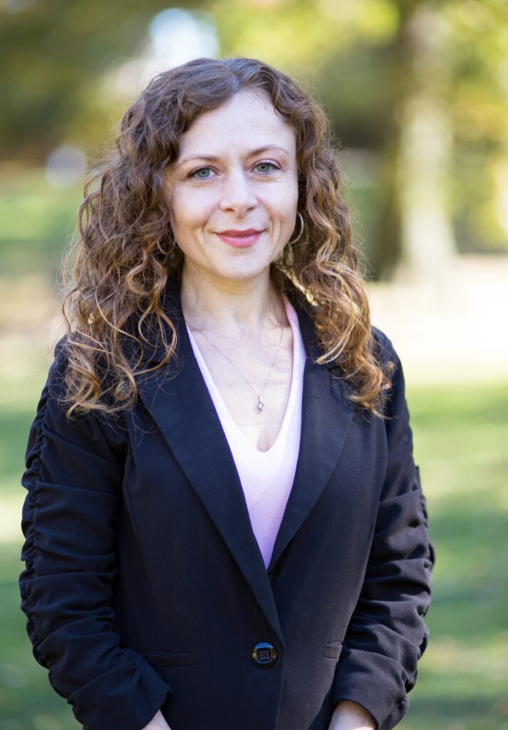 A woman with curly hair is standing outdoors, smiling at the camera. She is wearing a black blazer over a light pink top. The background shows greenery and sunlight, suggesting it is a park or garden.