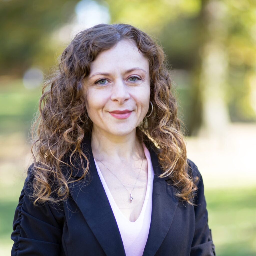 A woman with curly hair is standing outdoors, smiling at the camera. She is wearing a black blazer over a light pink top. The background shows greenery and sunlight, suggesting it is a park or garden.