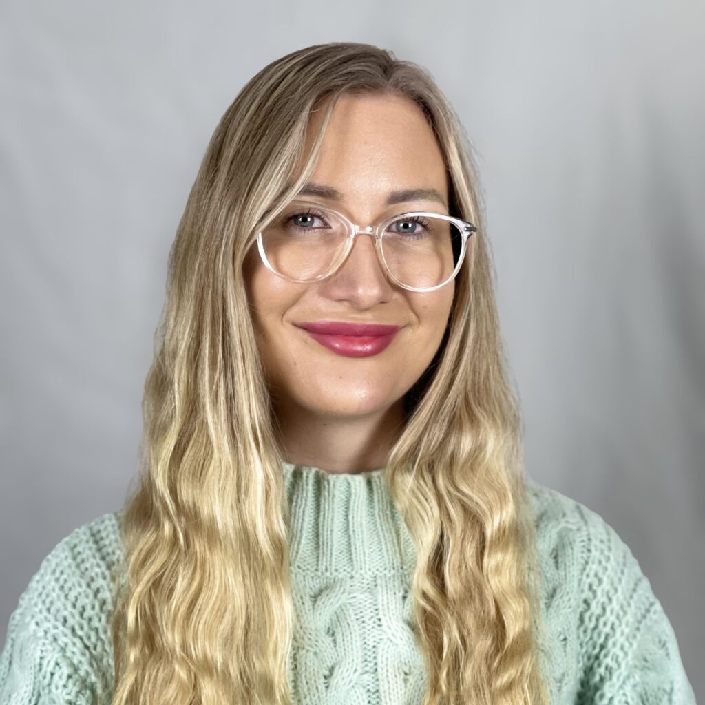Person with long blonde hair and glasses, wearing a light green knit sweater, smiles in front of a plain background.
