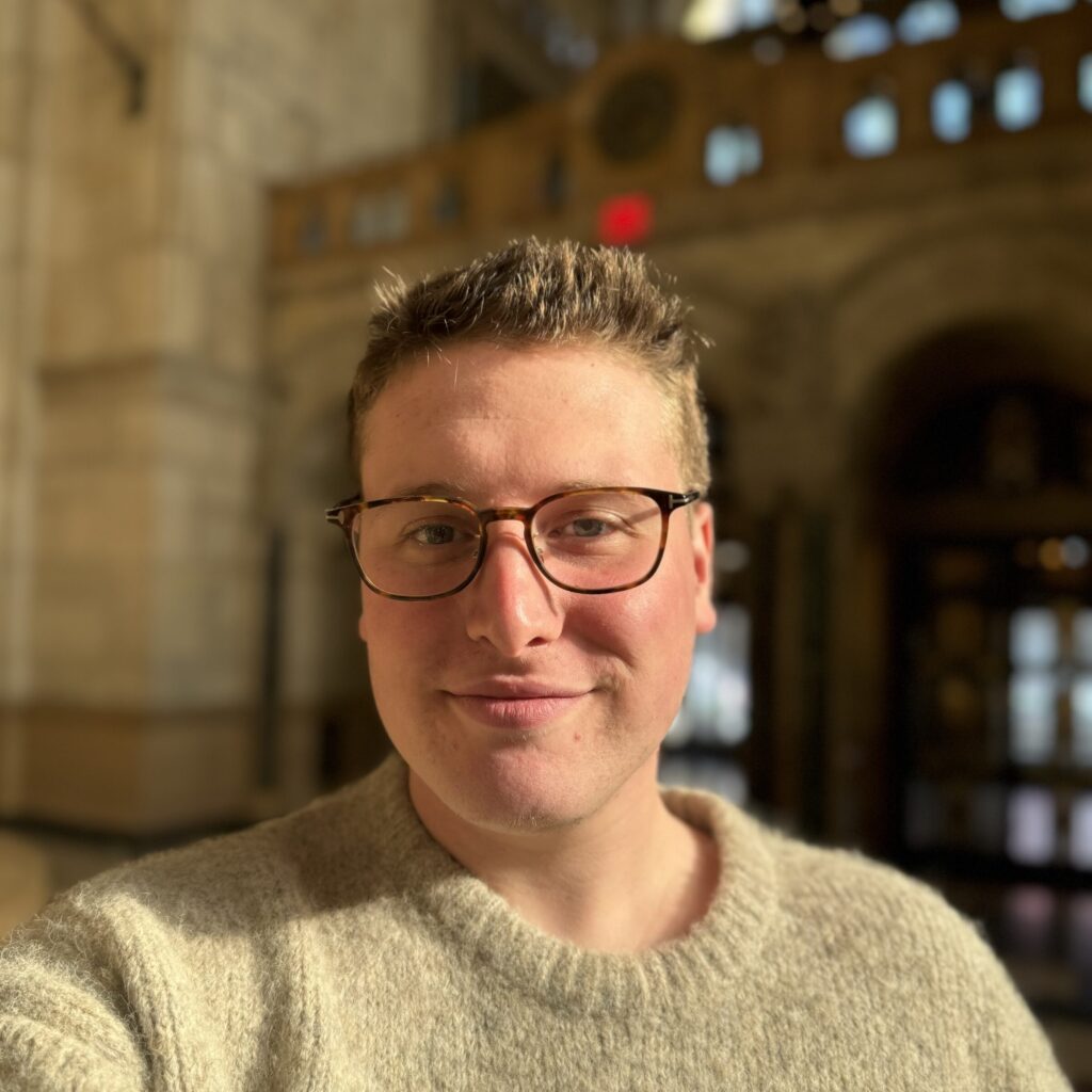 A person wearing glasses and a beige sweater smiles softly while taking a selfie in a well-lit, grand architectural interior with high ceilings and arched windows.