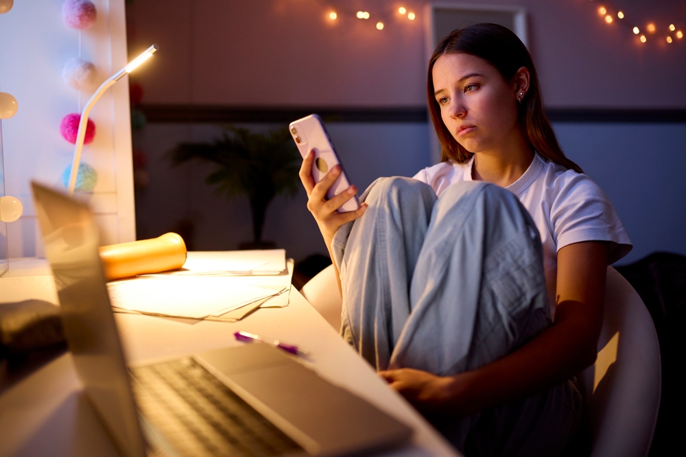 A teenage girl sits at a desk in a dimly lit room, looking at her phone with a serious expression. She has her knees pulled up to her chest and a laptop, papers, and a pen are on the desk in front of her.