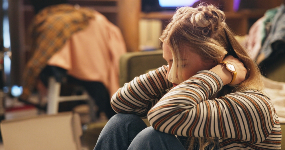A young person with long blonde hair and a striped shirt sits with their head resting on their knees, looking down. The background shows a cluttered room with clothing and furniture.