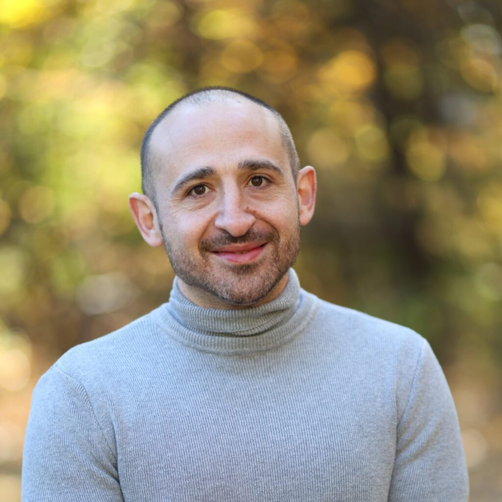 A man with short hair and a trimmed beard is standing outdoors, wearing a light gray turtleneck sweater and smiling softly. The background is blurred with warm, yellow-green tones of foliage.