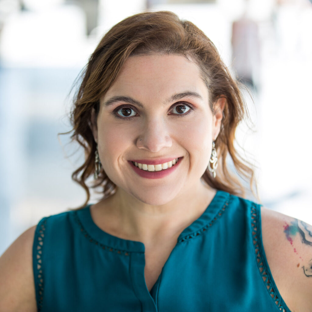 A woman with wavy, light brown hair and earrings smiles at the camera. She is wearing a sleeveless teal blouse and has a visible tattoo on her upper arm. The background is softly blurred.