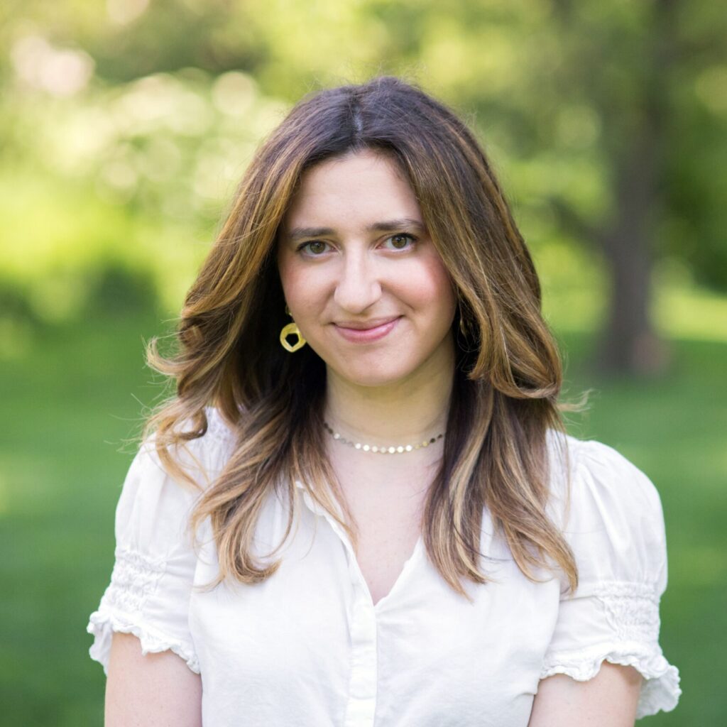 A woman with long brown hair, wearing a white blouse and gold hoop earrings, stands outside on a sunny day with green grass and trees in the background.