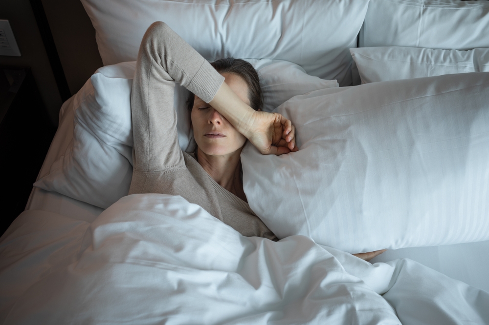A woman lies in bed under white blankets, resting her head on a pillow and covering her eyes with her arm, appearing tired or unwell. The setting is cozy and softly lit, suggesting morning or relaxation.