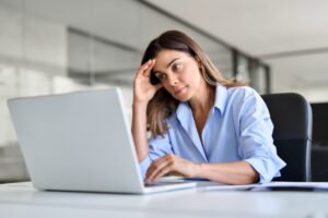 A woman in a blue shirt sits at a desk, looking concerned or stressed as she looks at her laptop screen with one hand on her forehead.