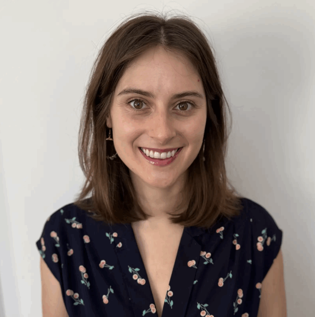 A woman with shoulder-length brown hair smiles at the camera, wearing a navy blouse with a pink floral pattern, standing against a plain white background.