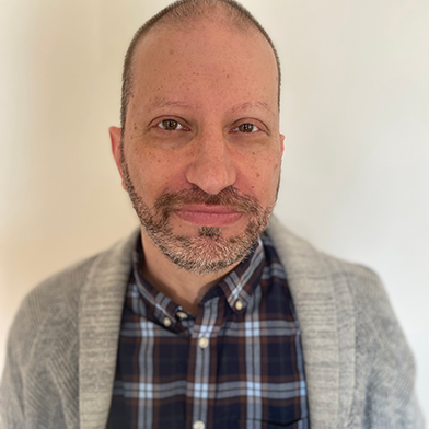 A man with a beard and mustache, wearing a gray sweater over a plaid shirt, stands in front of a plain light-colored background, looking at the camera with a neutral expression.