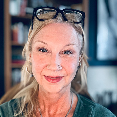 A middle-aged woman with blonde hair and glasses resting on her head smiles softly at the camera. She wears a hoop earring, a nose ring, and a green top, with bookshelves blurred in the background.