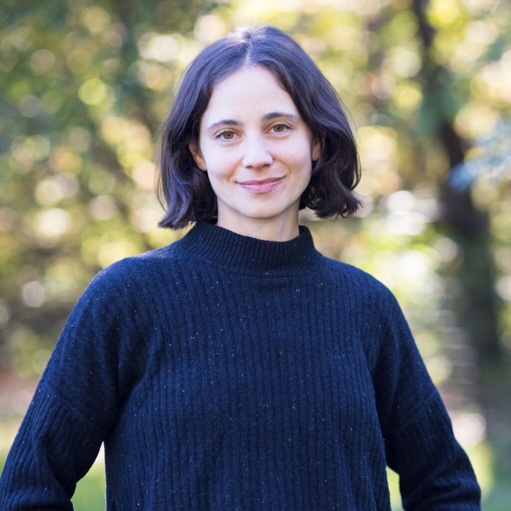 A woman with short dark hair wearing a black sweater stands outdoors, smiling softly. The background is blurred with green and yellow foliage, suggesting a park or natural setting.