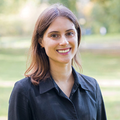 A young woman with straight brown hair and a friendly smile stands outdoors in a park, wearing a black button-up shirt. The background is blurred greenery.