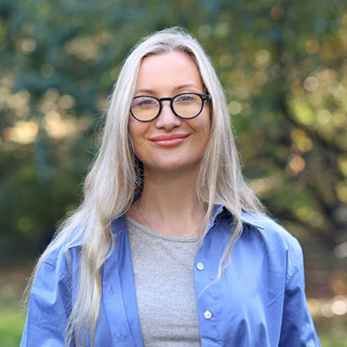 A woman with long blonde hair and glasses is smiling outdoors. She is wearing a light gray shirt under a blue button-up shirt, with a blurred background of green trees.