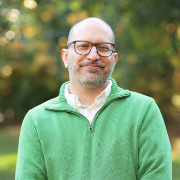 A man with glasses and a beard wears a green zip-up sweater over a white shirt, standing outdoors with a blurred green and yellow background.
