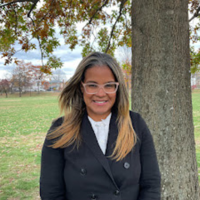 A woman with long, light brown hair and glasses stands outdoors in front of a tree, wearing a black coat and white shirt. Green grass and trees with autumn leaves are in the background.