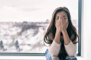 A woman sits indoors by a window, covering her face with her hands. She appears distressed or upset. The outside view shows a snowy, blurred landscape.