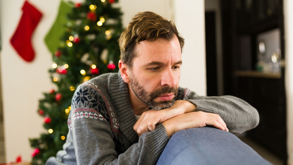 A man in a gray patterned sweater rests his arms and chin on the back of a couch, looking sad. A decorated Christmas tree and stockings are visible in the background.