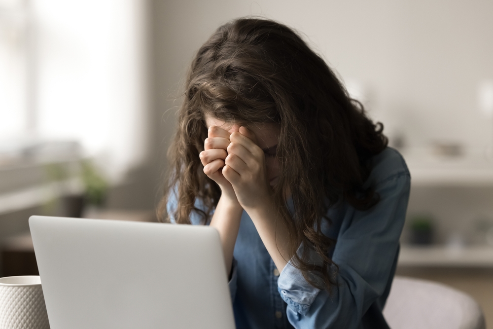 A person with long brown hair sits in front of a laptop, head bowed and hands covering their face, appearing stressed or upset. The background is softly blurred.