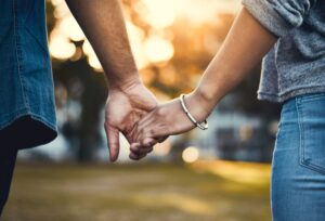 Close-up of two people holding hands outdoors, with warm sunlight in the background. One person wears a bracelet and a gray sleeve, the other a blue shirt. The scene conveys a sense of connection and affection.