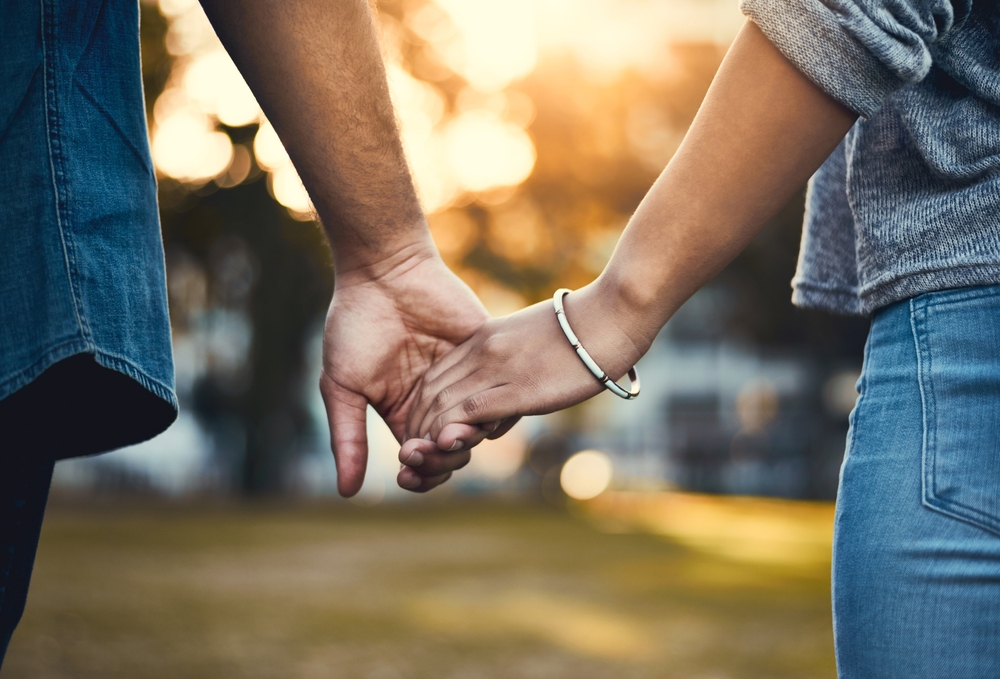 Close-up of two people holding hands outdoors, with warm sunlight in the background. One person wears a bracelet and a gray sleeve, the other a blue shirt. The scene conveys a sense of connection and affection.