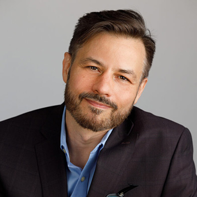 A man with short brown hair and a beard, wearing a dark blazer over a light blue shirt, smiles gently at the camera against a neutral background.
