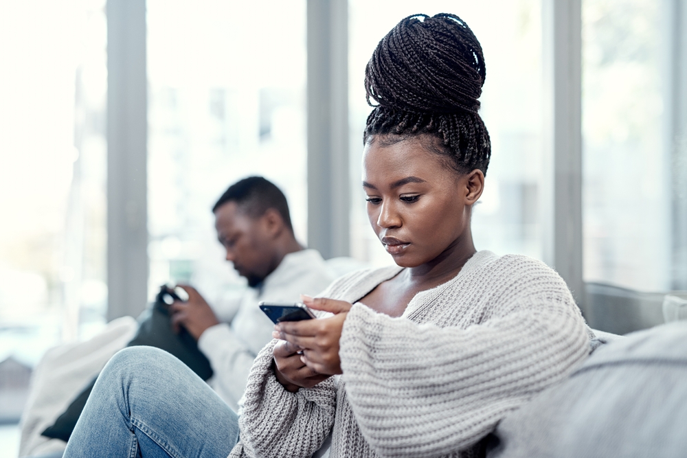 A woman with braided hair sits on a couch looking at her phone, appearing focused. In the background, a man also sits on the couch using his phone. Both are indoors with natural light coming through windows.
