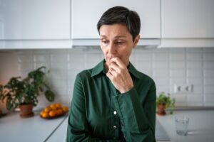 A woman with short dark hair wearing a green shirt stands in a kitchen, looking thoughtful and concerned with her hand touching her mouth. Potted plants and a bowl of oranges are visible in the background.