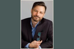 A man with short dark hair and a beard, wearing a dark suit jacket over a blue shirt, sits and smiles at the camera while holding eyeglasses in one hand. The background is plain and light-colored.