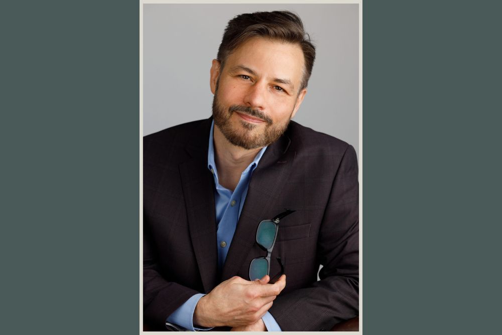 A man with short dark hair and a beard, wearing a dark suit jacket over a blue shirt, sits and smiles at the camera while holding eyeglasses in one hand. The background is plain and light-colored.