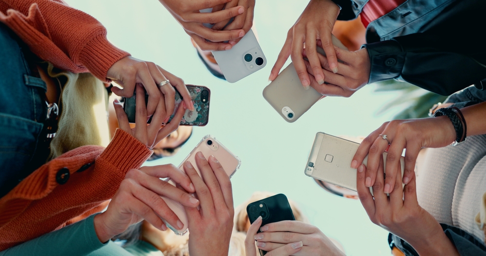 A group of people standing in a circle, holding and using smartphones, seen from below with a clear sky background. Multiple hands and phones are visible, suggesting social interaction or sharing.