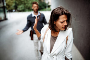 A woman in a white coat turns away, looking upset and holding up her hand to dismiss a man behind her, who appears frustrated, on a city street.
