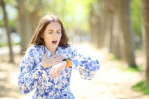 A woman in a blue floral dress yawns and looks at her watch while standing outdoors on a sunny day, appearing tired or sleepy. Trees and sunlight are blurred in the background.