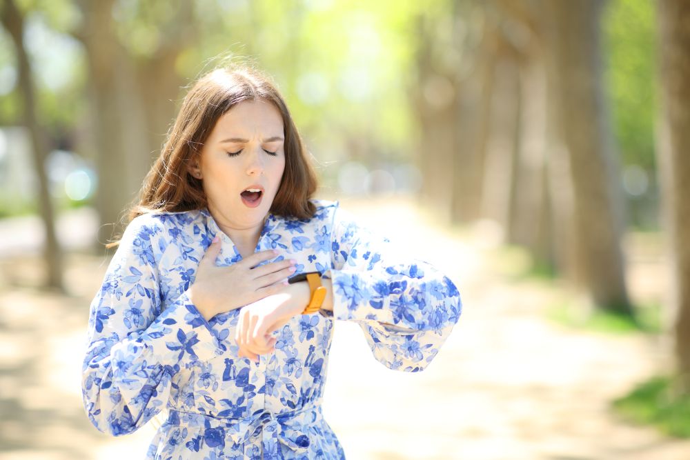 A woman in a blue floral dress yawns and looks at her watch while standing outdoors on a sunny day, appearing tired or sleepy. Trees and sunlight are blurred in the background.