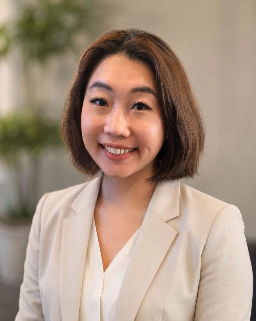 A woman with short brown hair wearing a beige blazer and white blouse smiles at the camera. The background is softly blurred with a green plant visible.