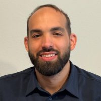 A man with a beard and short hair, wearing a dark button-up shirt, sits in a chair against a plain light-colored wall, smiling at the camera with his hands folded in his lap.