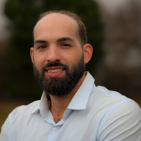 A bearded man with short hair, wearing a light blue collared shirt, stands outdoors with blurred trees and foliage in the background, smiling slightly at the camera.